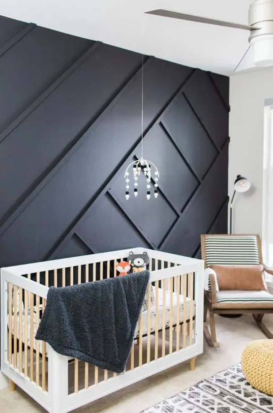 a bold nursery with a black geometric paneled wall, light-colored wooden furniture, geometric bedding and a cute mobile