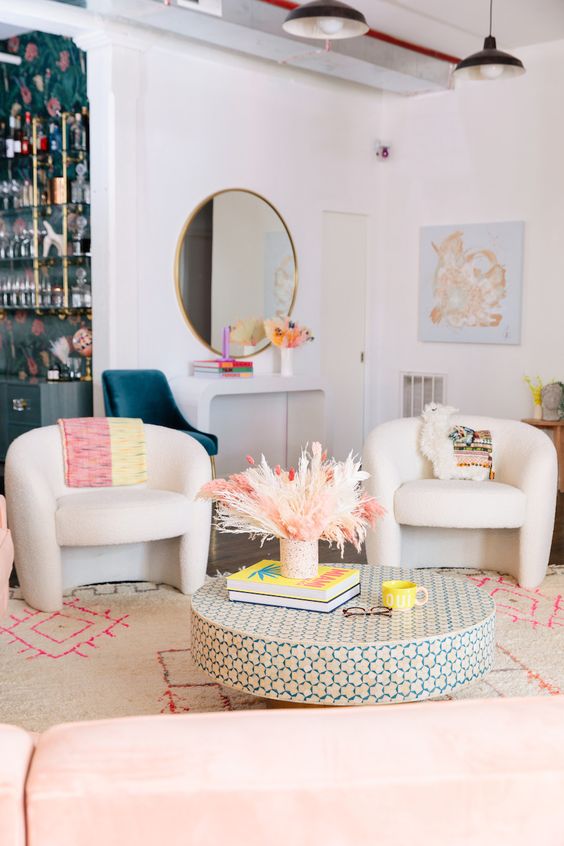 a bright and whimsy living room with a curved console and a round mirror, curved chairs, a round coffee table and pink touches