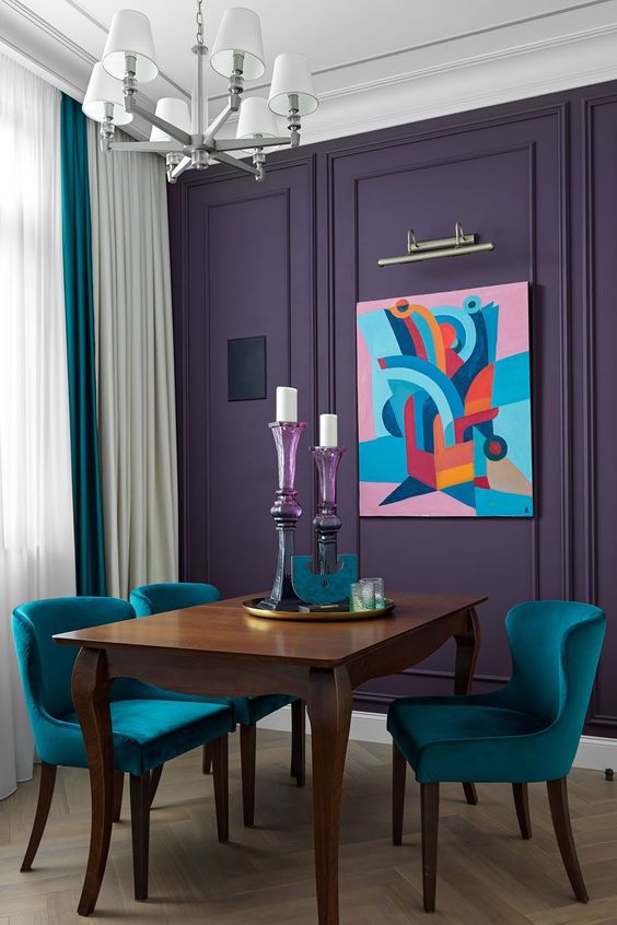 a catchy and bold dining room with a violet accent wall, a refined dining table, teal chairs, a chic chandelier and candleholders