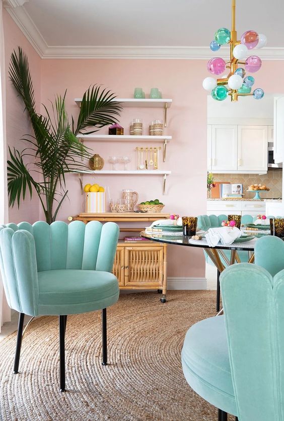 a catchy dining space with pink walls, a rattan storage unit, a glass top dining table and aqua chairs, a pastel bubble chandelier