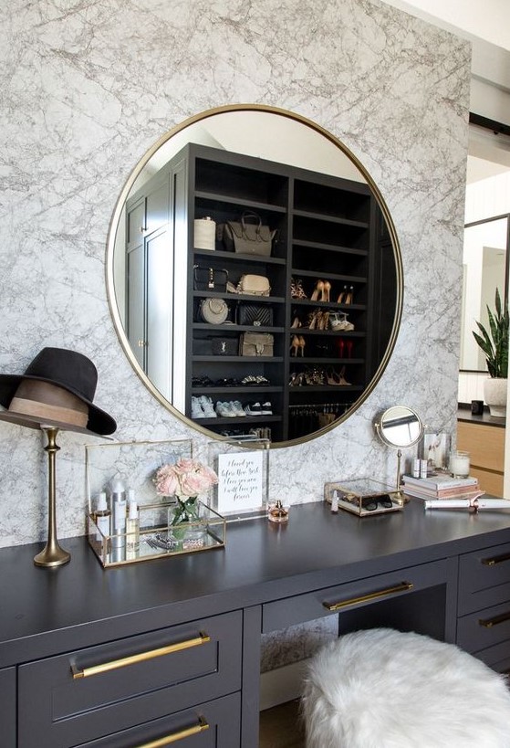 a chic closet with a makeup nook with a marble wall, a grey vanity, a gold frame mirror and many accessories
