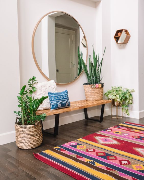 a colorful boho entryway with a stained bench, an oversized round mirror, a colorful printed rug and potted plants