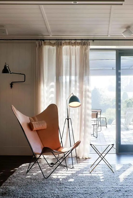 a cool nook by the window with an amber leather butterfly chair and a side table, a black floor lamp and a sconce