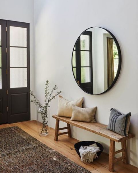 a cozy boho entryway with a round mirror in a black frame,a  stained bench with pillows and a printed rug
