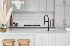 a dove grey kitchen with white skinny tiles on the backsplash and kitchen island, a macrame chandelier and wooden stools