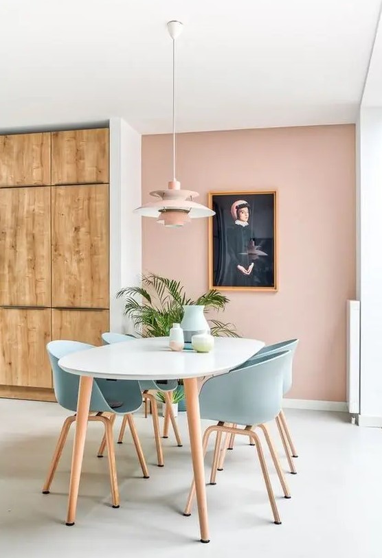 a dusty pink accent wall, a white dining table, pastel blue chairs, a pink pendant lamp, an artwork and a stained storage unit