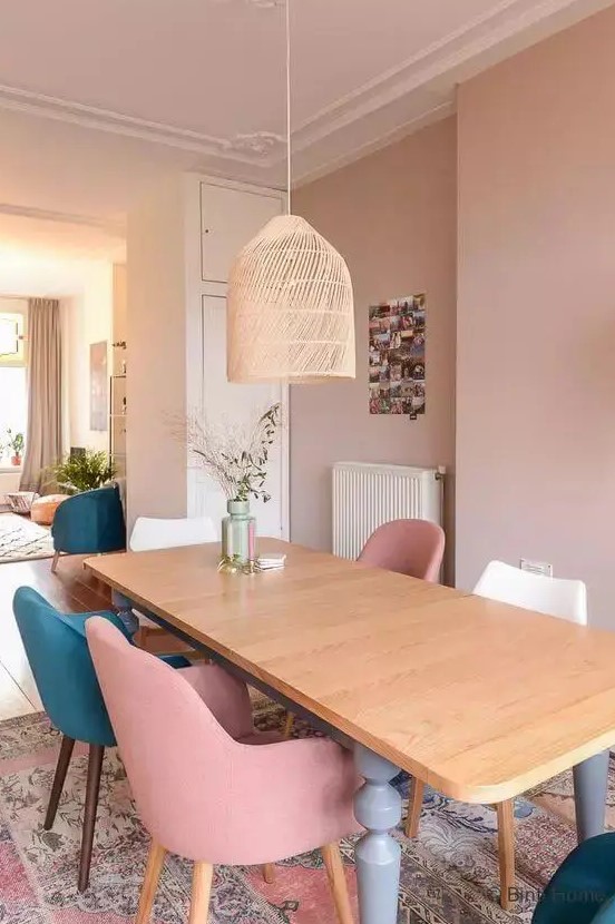 a dusty pink dining room with a stained table, dusty pink, white and teal chairs, a pink rug and a woven pendant lamp