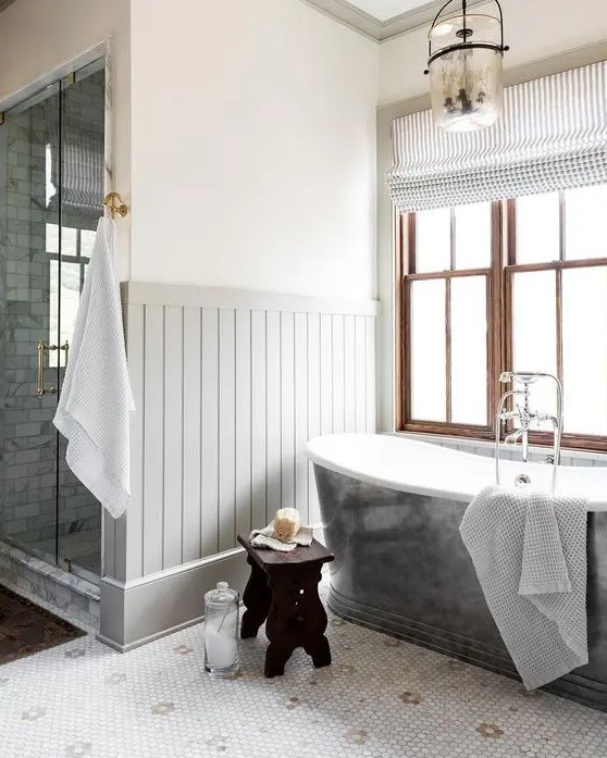 a farmhouse bathroom with paneling and penny tiles on the floor, a metal clad tub, stained double-hung windows, white towels