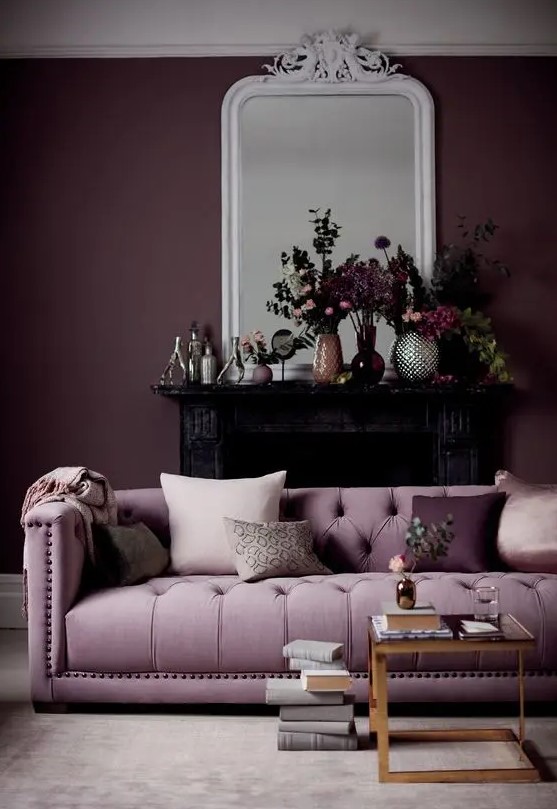 a gorgeous living room with a deep purple accent wall, a lilac sofa, a glass coffee table, a black fireplace and beautiful blooms on the mantel
