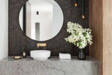 a gorgeous modern bathroom with a grey tile wall, black lamps, a grey stone vanity and a round mirror