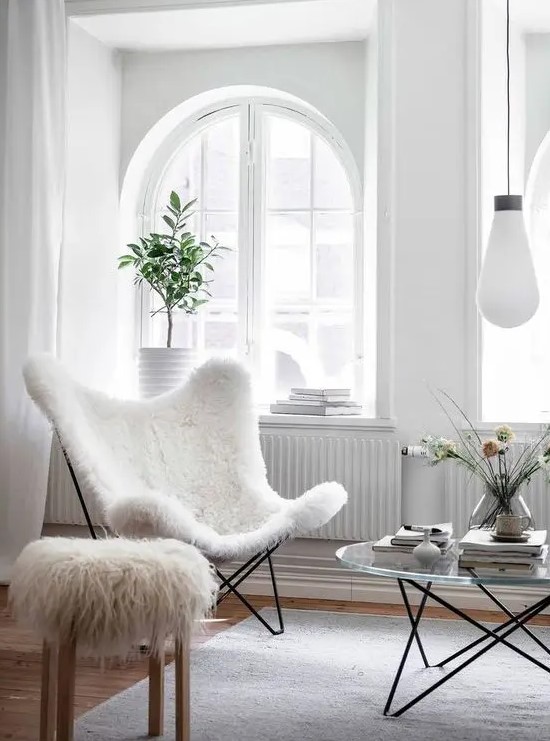 a light-filled living room with arched windows, a white faux fur butterfly chair, a faux fur stool and a glass coffee table