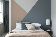 a lovely bedroom with a grey, rust and dark grey geometric accent wall, a bed with matching in color bedding and a white lamp