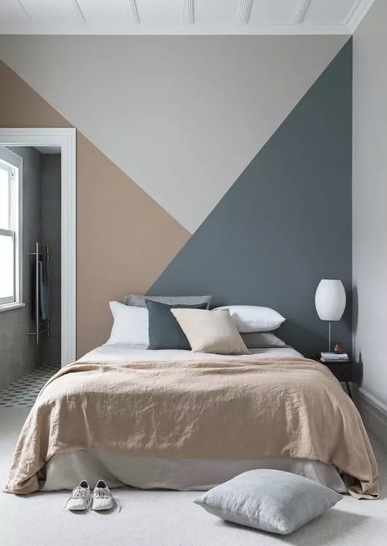 a lovely bedroom with a grey, rust and dark grey geometric accent wall, a bed with matching in color bedding and a white lamp