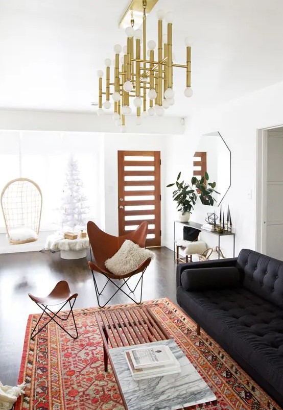 a lovely boho meets modern space with a black sofa, a tan leather butterfly chair and a footrest, a printed rug, a gilded chandelier
