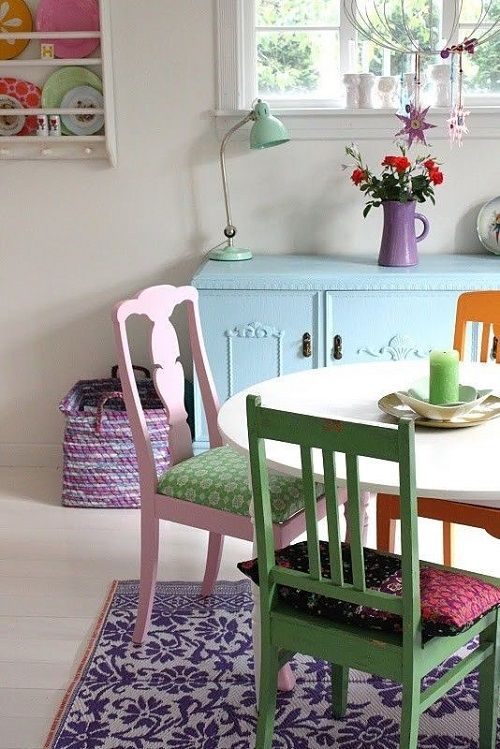 a lovely bright and pastel dining nook with a blue credenza, a round table, mismatching pastel chairs and a purple rug