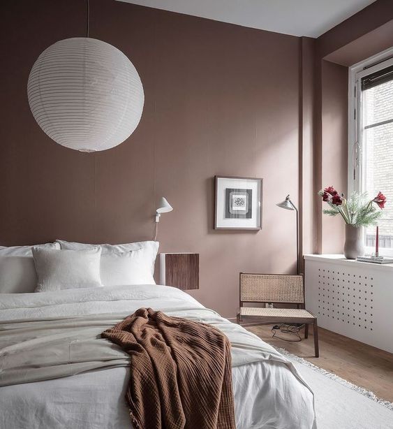 a mauve Scandinavian bedroom with a bed with white bedding, a sphere pendant lamp, a rattan chair and an artwork