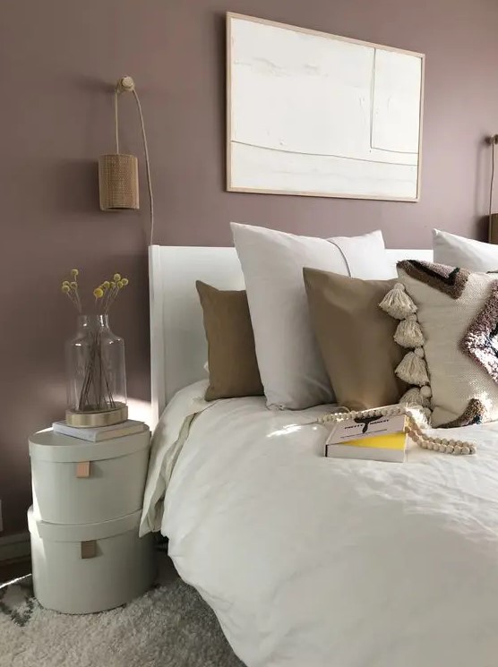 a mauve bedroom with a white bed with neutral boho bedding, stacked boxes, wooden sconces and a statement artwork