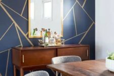 a mid-century modern dining room with a navy and gold geometric wall, a stained credenza and dining table, grey and brass chairs