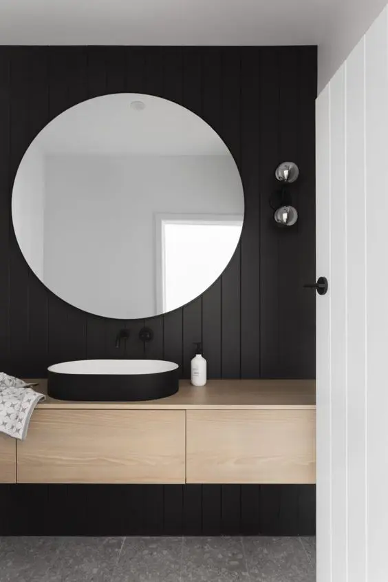 a minimalist bathroom with a black accent wall, a stained vanity, a black sink and a large round mirror to make the space look bigger