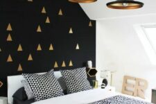 a modern and contrasting bedroom with a black and gold triangle accent wall, neutral furniture, black and white geometric print bedding and gold and black pendant lamps