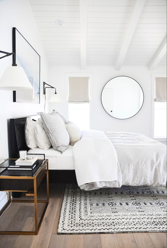 a modern bedroom with a black bed, neutral bedding, a printed rug, a black nightstand and a round mirror