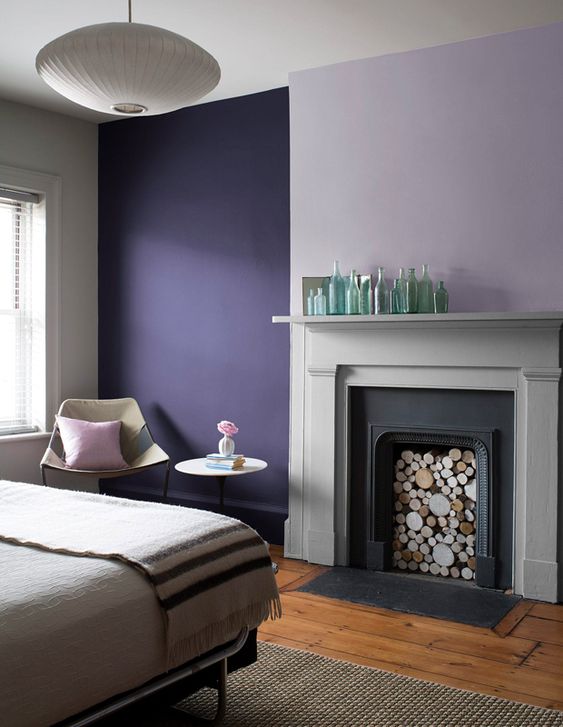 a modern bedroom with a color block accent wall done with violet and lavender, a non working fireplace, a bed with neutral bedding and a neutral chair