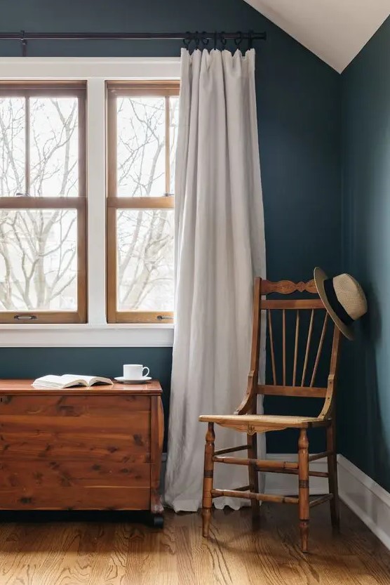 a modern farmhouse space with navy walls, a stained chair and chest and echoing stained double-hung windows and white curtains