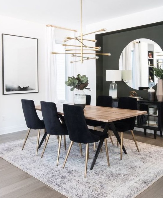 a modern glam dining room with a grey accent wall, a large round mirror, a catchy chandelier and black chairs