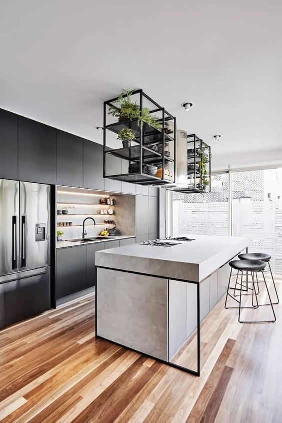 a modern industrial kitchen in black, with built-in shelves, a concrete kitchen island, suspended shelves over it and black stools