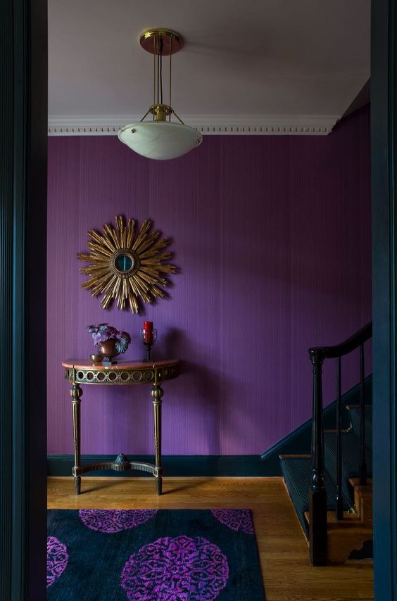 a moody and sophisticated entryway with a purple accent wall, a refined carved console table, a burst mirror and a printed rug