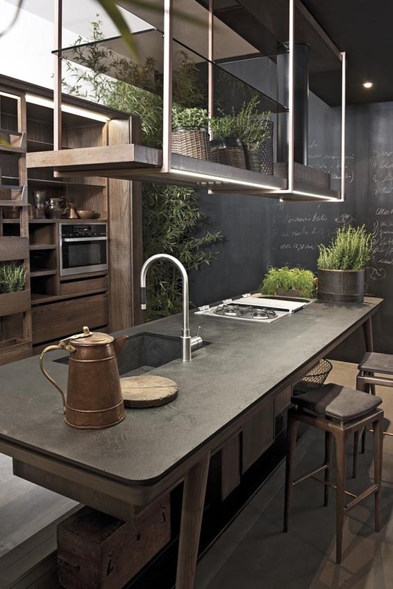 a moody industrial kitchen with stained cabinets, a table-style kitchen island and suspended shelves over this island, a chalkboard wall