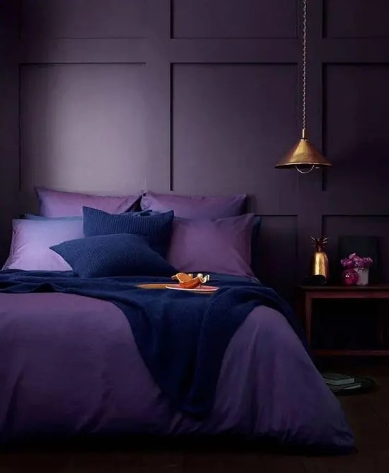 a moody monochromatic bedroom with deep purple paneled walls, purple and navy bedding, a wooden nightstand and a metal pendant lamp