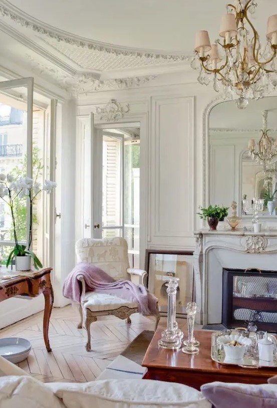 a neutral Parisian living room with a fireplace clad with marble, an antique neutral chair, some antique tables and a crystal chandelier