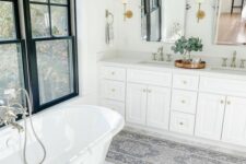 a neutral farmhouse bathroom with black-framed double-hung windows, a white vanity, white sinks, an oval vintage tub and a printed rug
