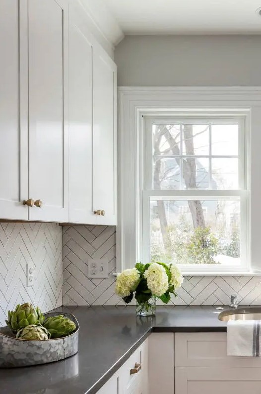 a neutral farmhouse kitchen with shaker cabinets, black countertops, a white herringbone tile backsplash and a small double-hung window
