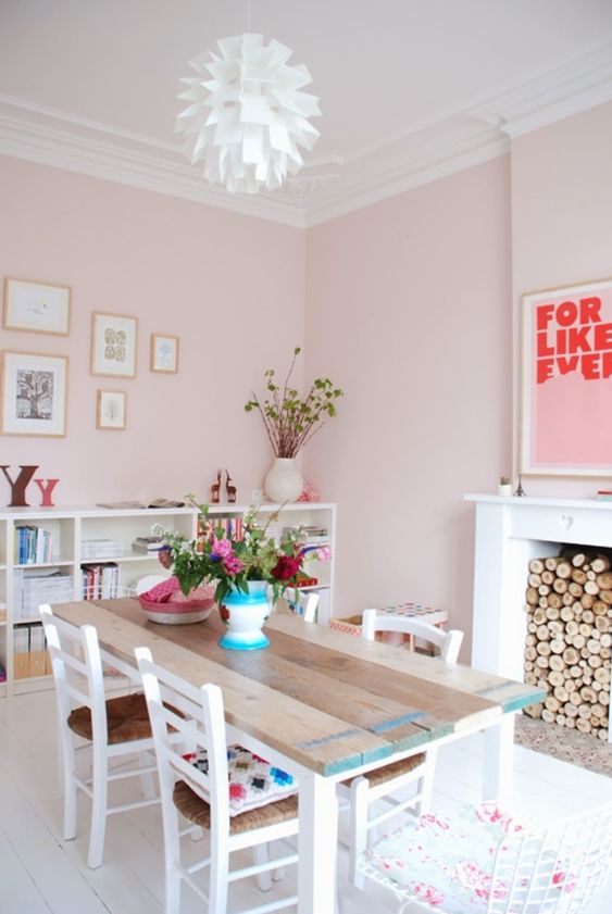 a pasle pink dining space with a non-working fireplace with firewood, a reclaimed wood table, matching chairs and a storage unit