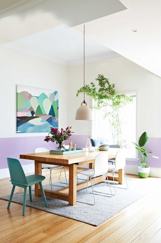 a pastel dining room with color block walls, a stained table, white and a green chair, a bold artwork and some potted plants