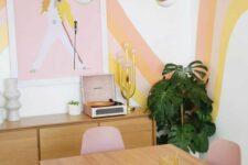 a pastel dining space with orange, pink and yellow patterns on the walls, an artwork, stained mid-century modern furniture and pink chairs