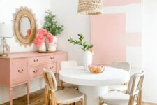 a pastel pink dining space with an accent wall, a pink credenza, a round table and neutral rattan chairs plus a woven pendant lamp