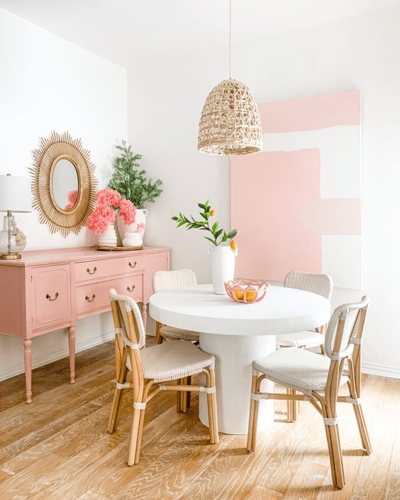 a pastel pink dining space with an accent wall, a pink credenza, a round table and neutral rattan chairs plus a woven pendant lamp