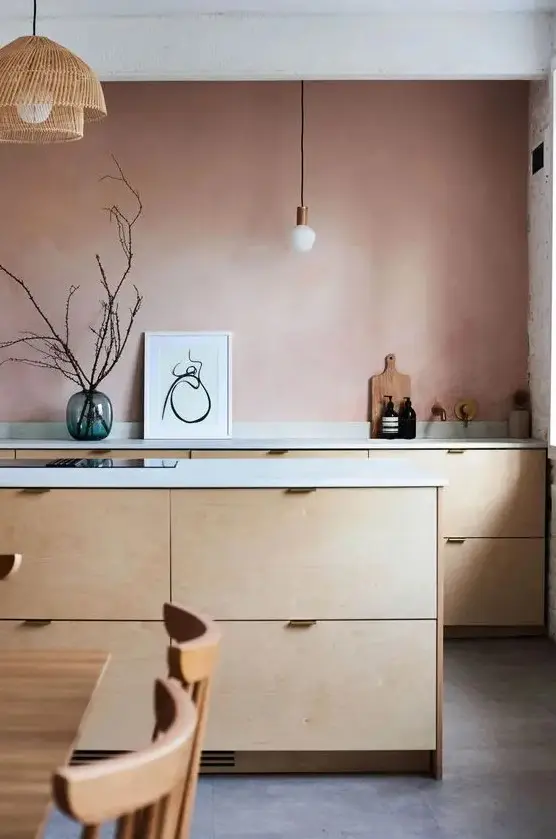 a pretty kitchen with a mauve accent wall, plywood cabinets, white countertops and pendant lamps
