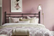 a pretty mauve bedroom with a black forged bed, neutral and pastel bedding, a stool, a sconce and a cool artwork