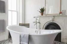 a refined and chic bathroom with a vintage French fireplace with a stone mantel, a free-standing tub, some art and beautiful tiles on the floor