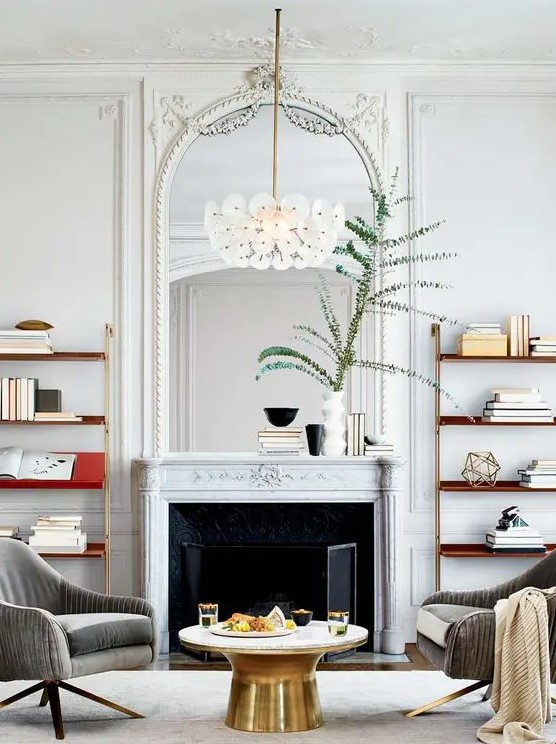 a refined living room with a French fireplace and an arched mirror in an ornated frame, bookshelves, a round coffee table and grey chairs
