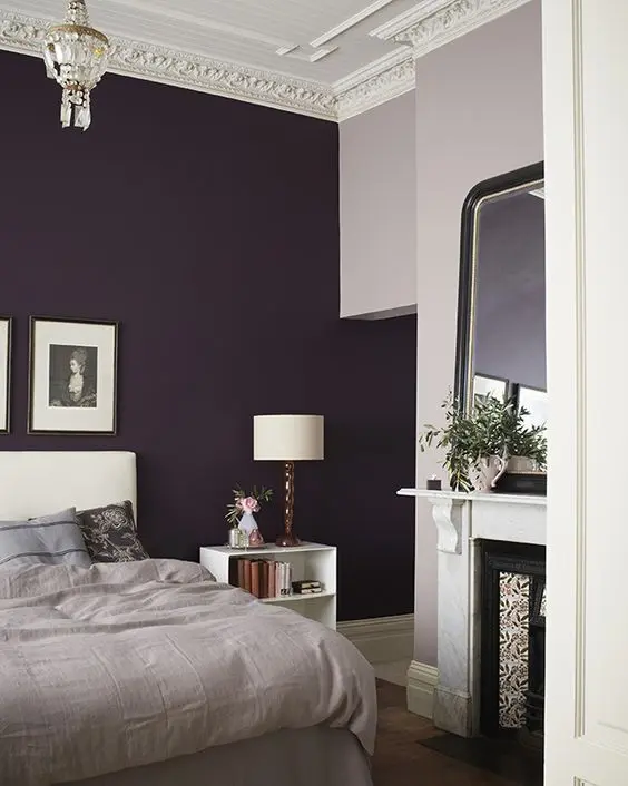 a refined moody bedroom with a deep purple accent wall, a creamy bed with neutral bedding, a non working fireplace, a white nightstand and some lamps