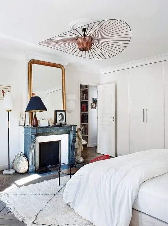 a serene Parisian bedroom with built-in wardrobes, a French fireplace with a blue mantel, a large mirror in a gilded frame and a bed with neutral bedding