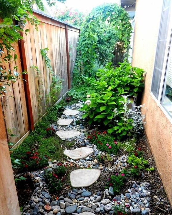 a small and bright garden nook with pebbles, greenery and bright blooms, shrubs and a climbing plants on the fence