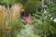 a small and textural garden with greenery, grasses, some trees and red metal furniture that is very well seen in tall grass