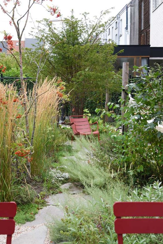 a small and textural garden with greenery, grasses, some trees and red metal furniture that is very well seen in tall grass