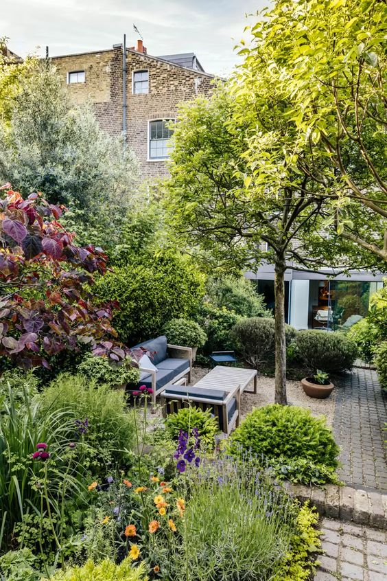 a small but very lush garden with greenery and bold blooms, shrubs and trees, neutral garden furniture and potted plants